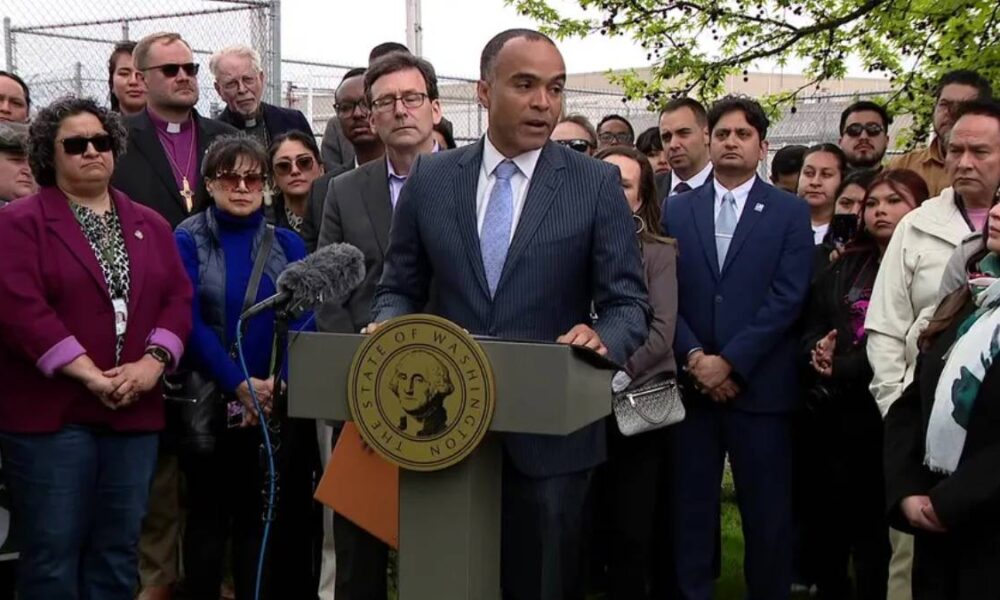 Man in a dark suit and light blue tie speaks at a podium with a microphone, surrounded by a diverse group of supporters and reporters.