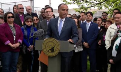 Man in a dark suit and light blue tie speaks at a podium with a microphone, surrounded by a diverse group of supporters and reporters.