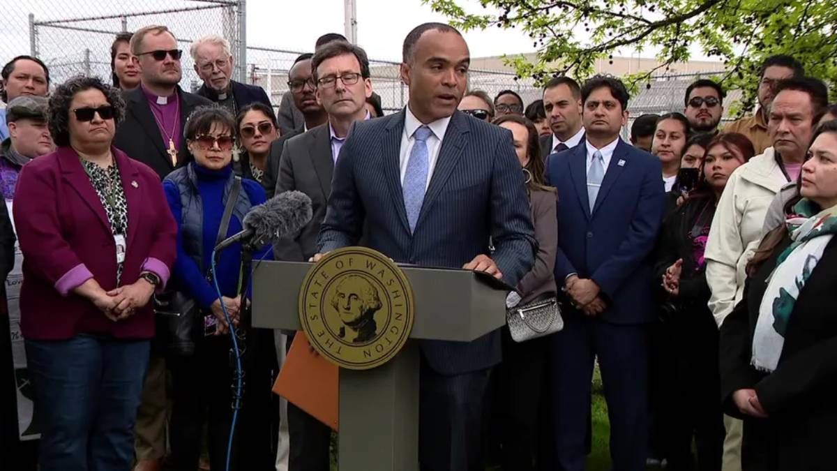 Man in a dark suit and light blue tie speaks at a podium with a microphone, surrounded by a diverse group of supporters and reporters.