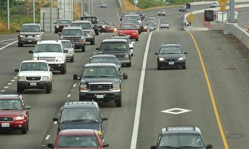 Heavy traffic on a multi-lane highway with many cars in the left lanes and a construction zone on the right shoulder barrier.
