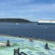 Bicyclists ride on a colorful mural-covered waterfront plaza with a large ship in the calm sea beyond and a forested shoreline in the distance.