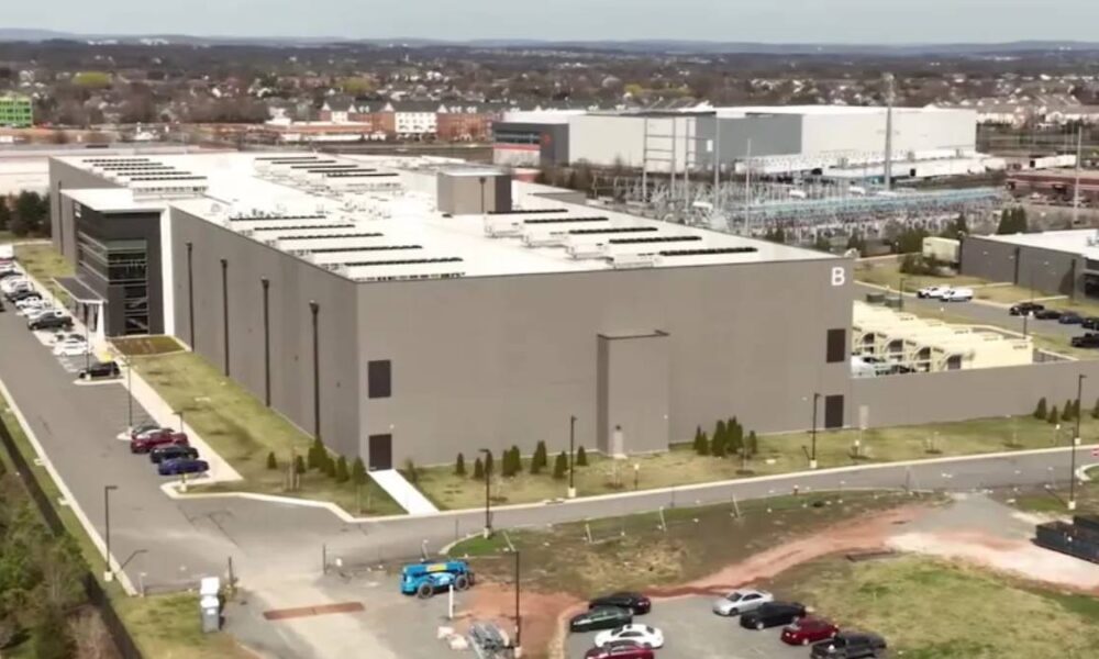 Aerial view of a large gray industrial building complex with a 'B' on the side, parking lots, and surrounding roads.