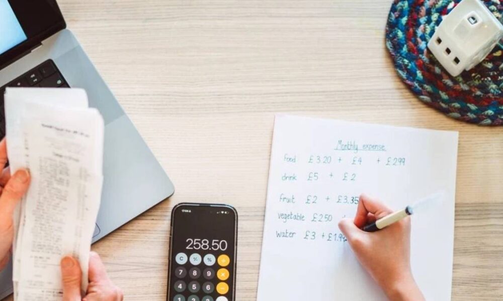 Desk scene with receipts, laptop, a calculator showing 258.50, and a person writing a monthly expenses list on paper.