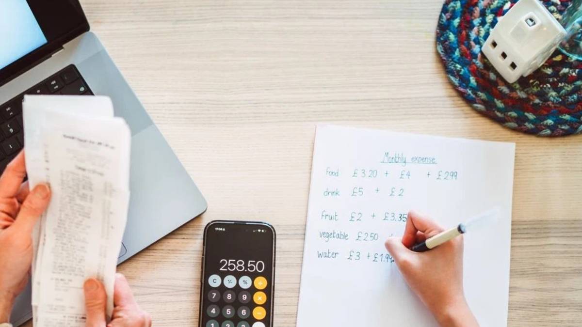 Desk scene with receipts, laptop, a calculator showing 258.50, and a person writing a monthly expenses list on paper.