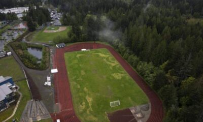 Aerial view of a red running track circling a grassy field with a goalpost, bordered by dense trees on one side and facilities on the other.