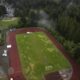 Aerial view of a red running track circling a grassy field with a goalpost, bordered by dense trees on one side and facilities on the other.