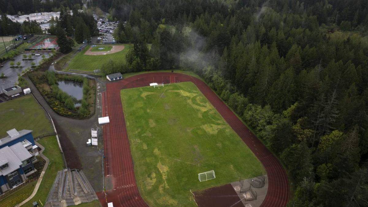 Aerial view of a red running track circling a grassy field with a goalpost, bordered by dense trees on one side and facilities on the other.