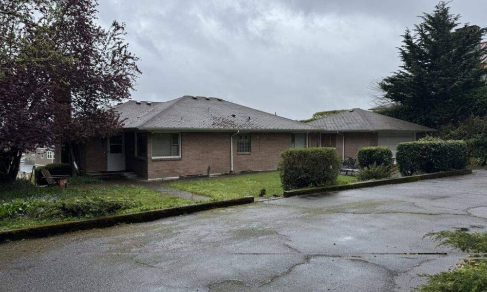 Single-story brick building with a sloped roof, shrubs in front, and a wet parking lot under a cloudy sky