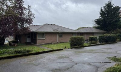 Single-story brick building with a sloped roof, shrubs in front, and a wet parking lot under a cloudy sky