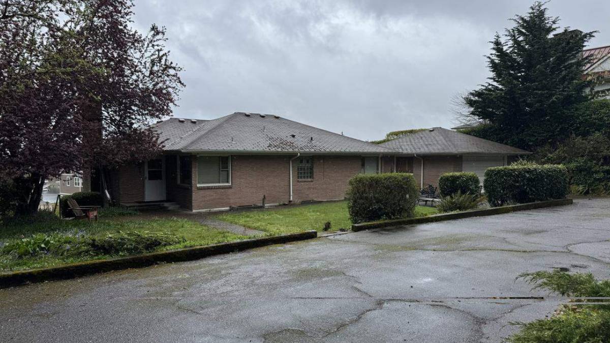 Single-story brick building with a sloped roof, shrubs in front, and a wet parking lot under a cloudy sky