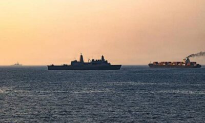 Silhouetted naval vessel and a container ship on calm sea at sunset.