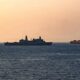Silhouetted naval vessel and a container ship on calm sea at sunset.
