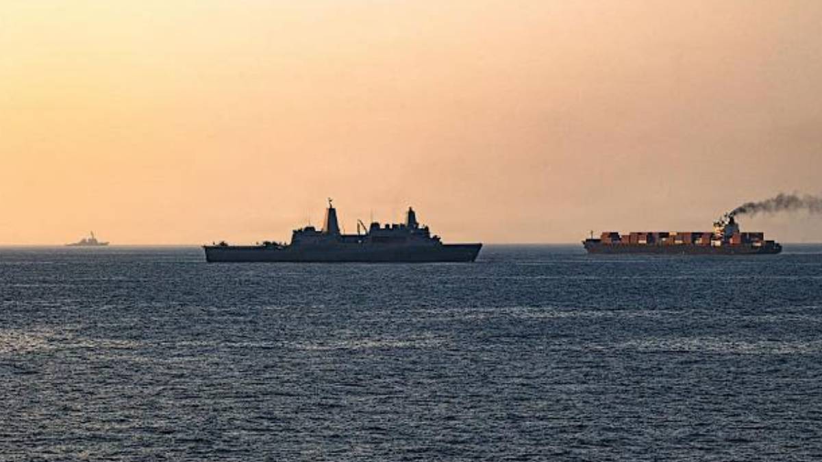 Silhouetted naval vessel and a container ship on calm sea at sunset.