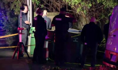 Nighttime crime scene with multiple investigators and police, yellow caution tape, and a van nearby under blue and purple lights.