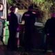 Nighttime crime scene with multiple investigators and police, yellow caution tape, and a van nearby under blue and purple lights.
