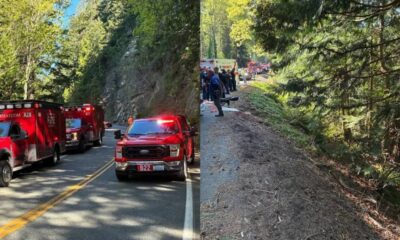 Emergency response on a rural road with red fire trucks and flashing lights blocking the lane; forested hillside in the background.
