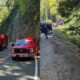 Emergency response on a rural road with red fire trucks and flashing lights blocking the lane; forested hillside in the background.
