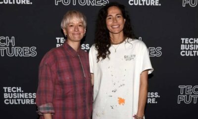 Two women stand side by side and smile at a tech event backdrop that says 'Tech. Business. Culture.' and 'FUTURES.' One wears a pink plaid shirt; the other wears a white paint-splattered T-shirt.