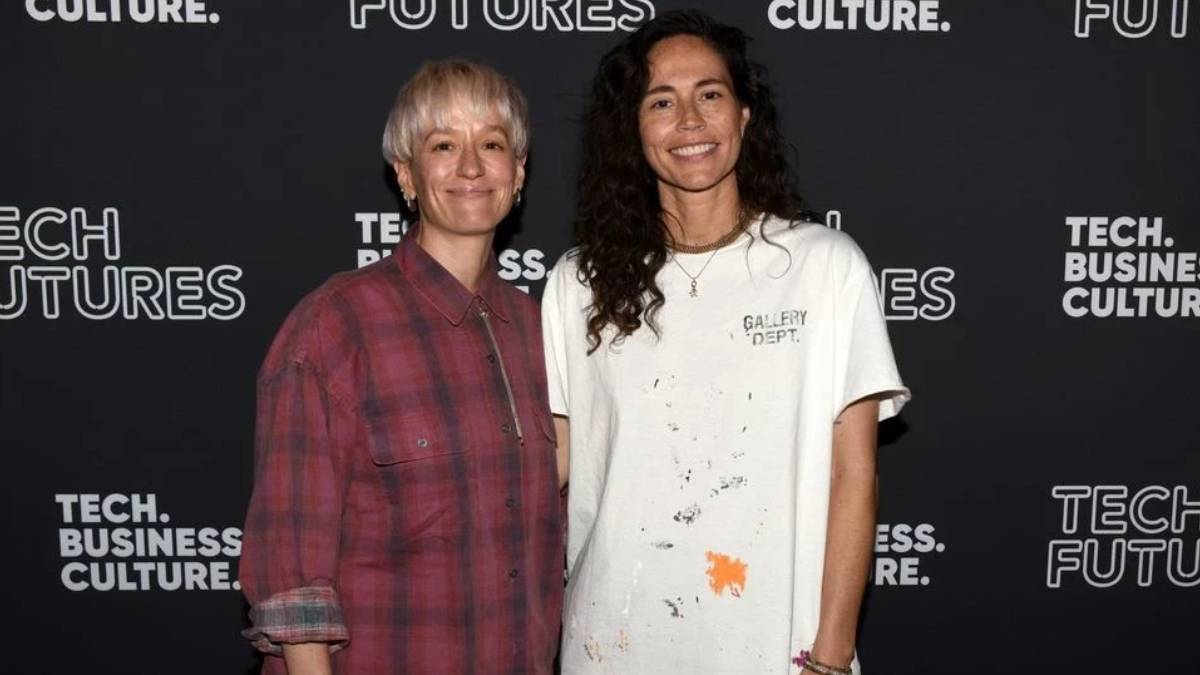 Two women stand side by side and smile at a tech event backdrop that says 'Tech. Business. Culture.' and 'FUTURES.' One wears a pink plaid shirt; the other wears a white paint-splattered T-shirt.