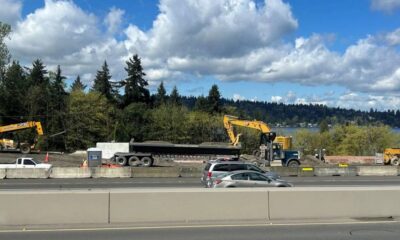 Construction site along a highway with yellow excavators, a long trailer and concrete barriers; trees and a lake in the background under a partly cloudy sky.