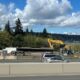 Construction site along a highway with yellow excavators, a long trailer and concrete barriers; trees and a lake in the background under a partly cloudy sky.