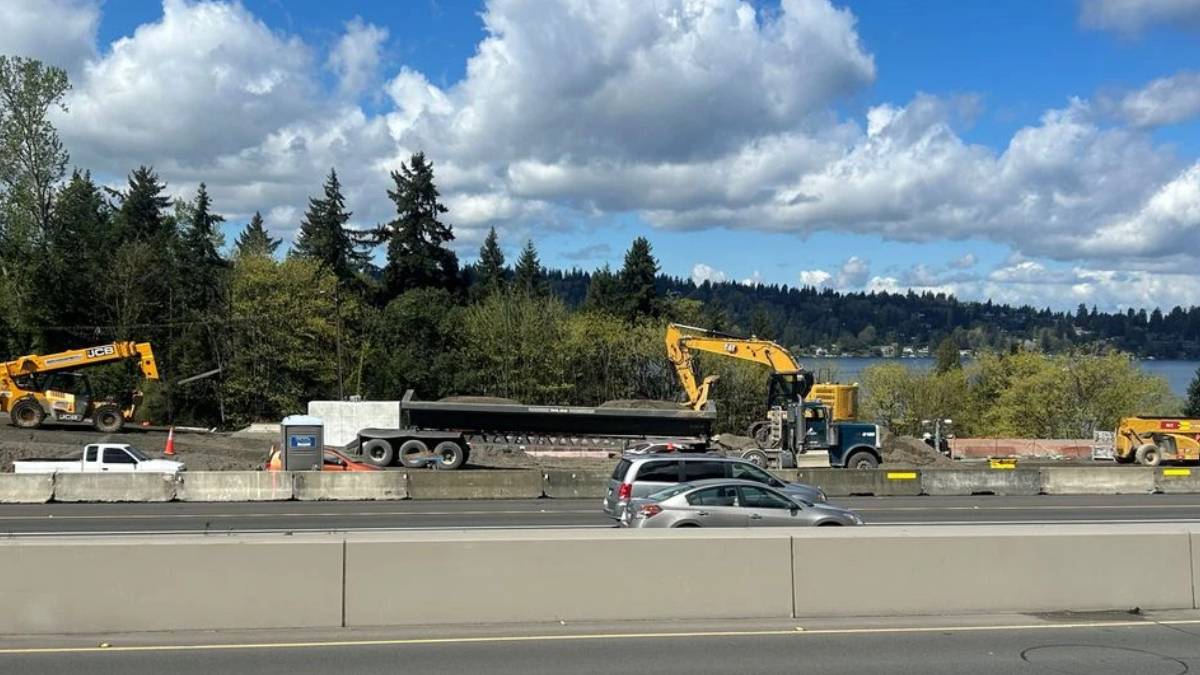 Construction site along a highway with yellow excavators, a long trailer and concrete barriers; trees and a lake in the background under a partly cloudy sky.