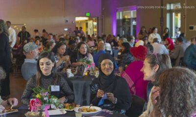 Diverse crowd at a large community dinner, people chatting and eating at round tables; foreground shows three women, one in a hijab, smiling.