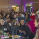 Diverse crowd at a large community dinner, people chatting and eating at round tables; foreground shows three women, one in a hijab, smiling.
