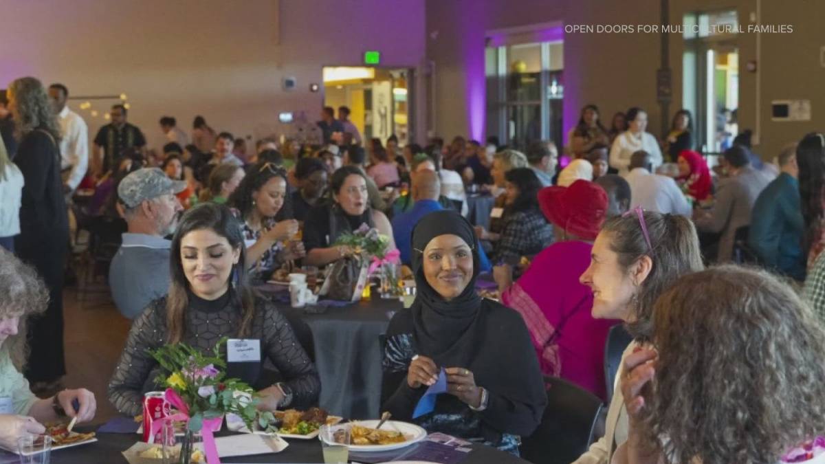 Diverse crowd at a large community dinner, people chatting and eating at round tables; foreground shows three women, one in a hijab, smiling.