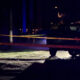 Night scene at a crime scene: yellow caution tape blocks a street with a dark SUV and faint emergency lights in the background.