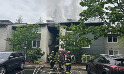 Firefighters in full gear fighting a blaze at a two‑story apartment building; smoke rising from the roof with hoses on the wet pavement in front.