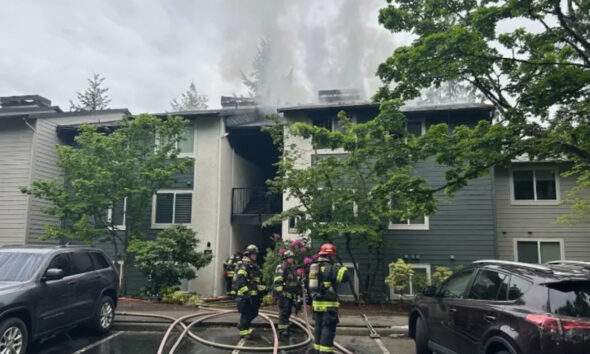 Firefighters in full gear fighting a blaze at a two‑story apartment building; smoke rising from the roof with hoses on the wet pavement in front.