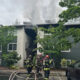 Firefighters in full gear fighting a blaze at a two‑story apartment building; smoke rising from the roof with hoses on the wet pavement in front.