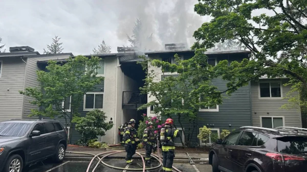 Firefighters in full gear fighting a blaze at a two‑story apartment building; smoke rising from the roof with hoses on the wet pavement in front.