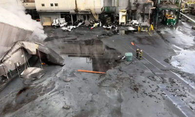 Industrial facility exterior with smoke rising from a damaged entryway; wet, charred ground and scattered debris as firefighters work nearby.