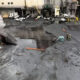 Industrial facility exterior with smoke rising from a damaged entryway; wet, charred ground and scattered debris as firefighters work nearby.