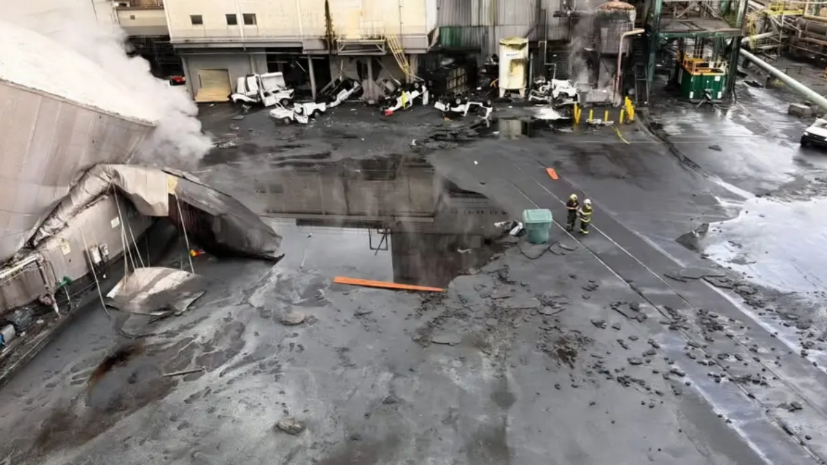 Industrial facility exterior with smoke rising from a damaged entryway; wet, charred ground and scattered debris as firefighters work nearby.