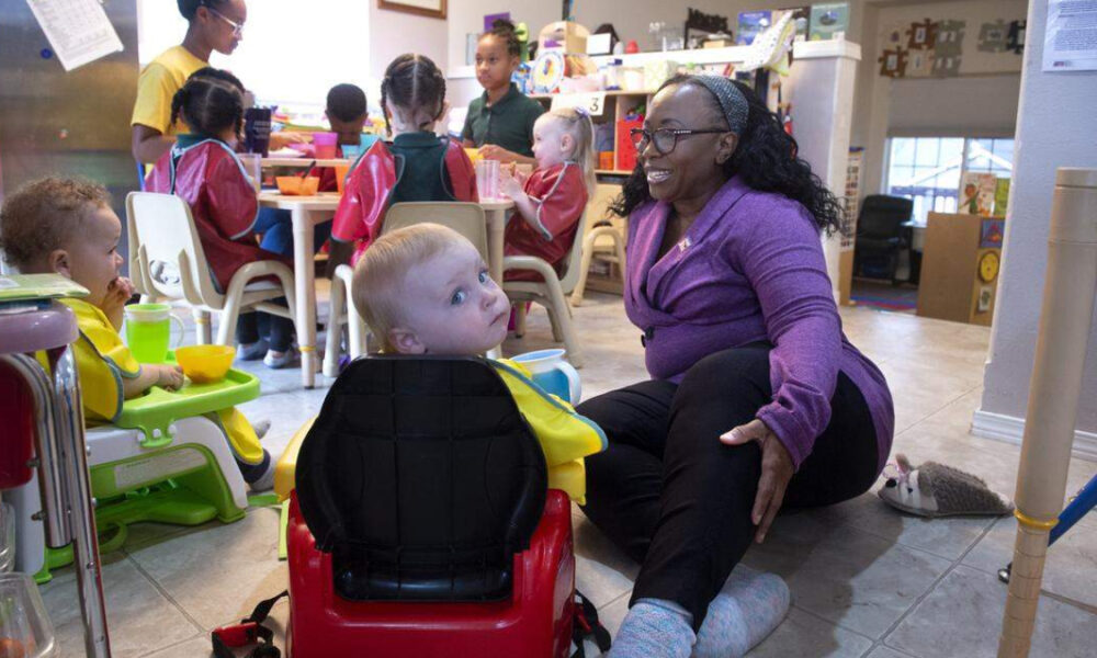Caregiver in a purple cardigan kneels beside a toddler in a red ride-on toy car, in a busy preschool classroom with other children at a table.
