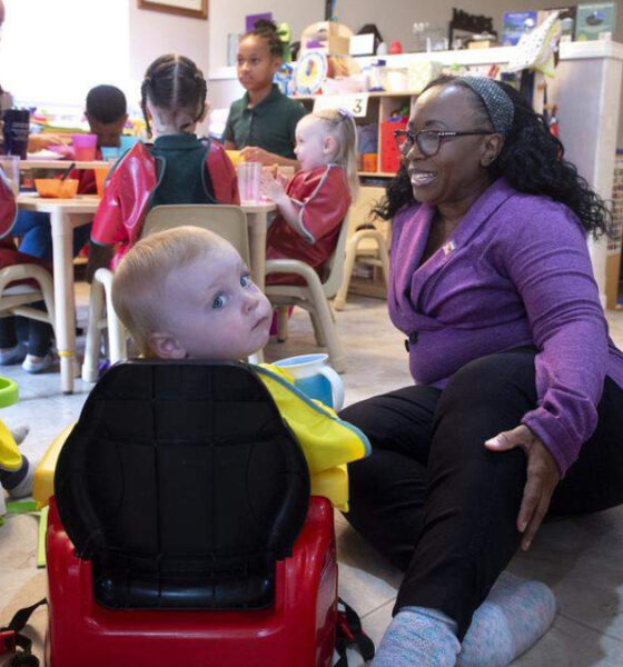 Caregiver in a purple cardigan kneels beside a toddler in a red ride-on toy car, in a busy preschool classroom with other children at a table.