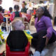 Caregiver in a purple cardigan kneels beside a toddler in a red ride-on toy car, in a busy preschool classroom with other children at a table.
