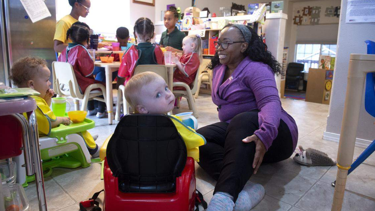 Caregiver in a purple cardigan kneels beside a toddler in a red ride-on toy car, in a busy preschool classroom with other children at a table.