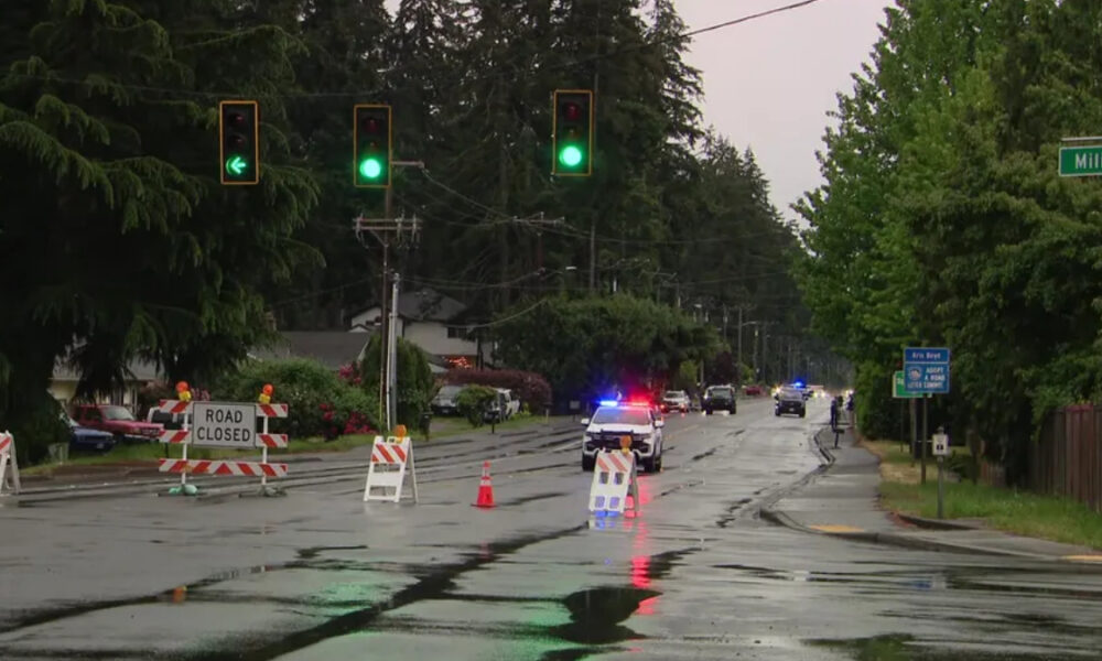 Closed suburban street with road-closure barricades and orange cones; a police car with flashing lights on a wet, reflective road corridor.