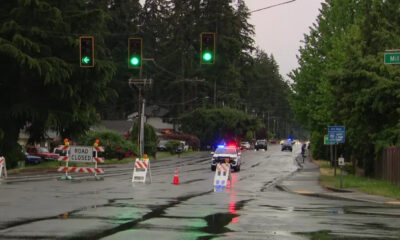Closed suburban street with road-closure barricades and orange cones; a police car with flashing lights on a wet, reflective road corridor.