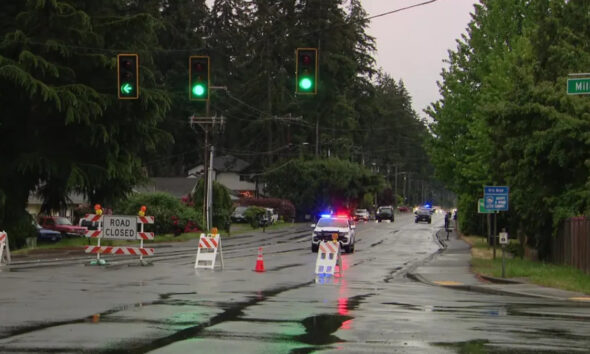 Closed suburban street with road-closure barricades and orange cones; a police car with flashing lights on a wet, reflective road corridor.