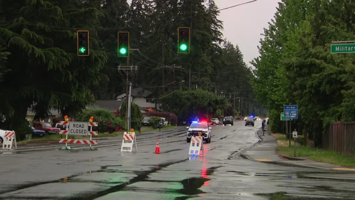 Closed suburban street with road-closure barricades and orange cones; a police car with flashing lights on a wet, reflective road corridor.