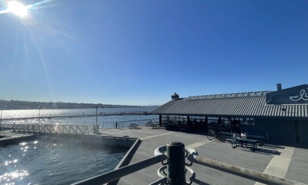 Sunlit waterfront scene with a blue sky, calm harbor, and a dockside restaurant building along the water. A pier and outdoor seating are visible.