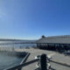 Sunlit waterfront scene with a blue sky, calm harbor, and a dockside restaurant building along the water. A pier and outdoor seating are visible.