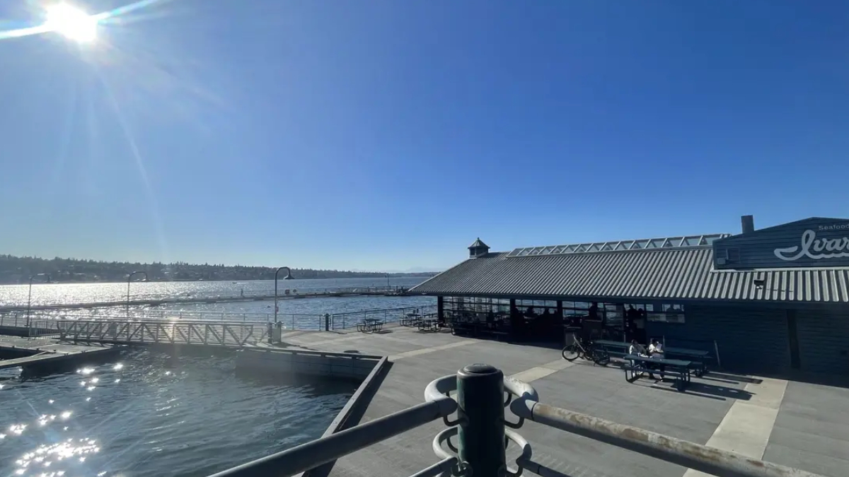 Sunlit waterfront scene with a blue sky, calm harbor, and a dockside restaurant building along the water. A pier and outdoor seating are visible.