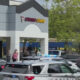 Police cars with flashing lights block a shopping center lot in front of a storefront, with pedestrians nearby.