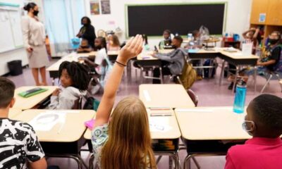 Student raises hand in a classroom as a teacher stands at the front.